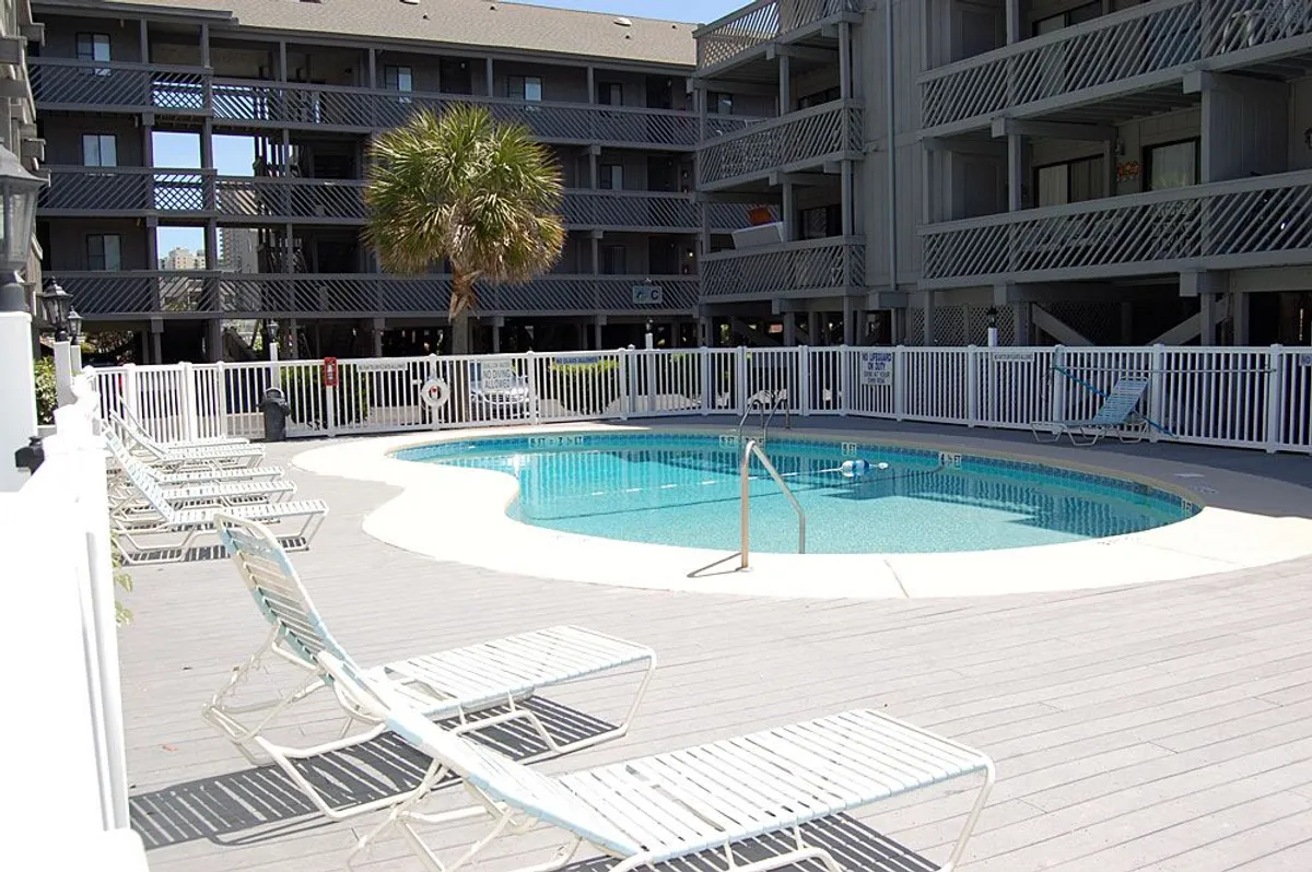 A sparkling outdoor swimming pool surrounded by palm trees and lounge chairs, enclosed by a fence for privacy. The setting is relaxed, perfect for unwinding after a day at the beach or enjoying family time by the water.