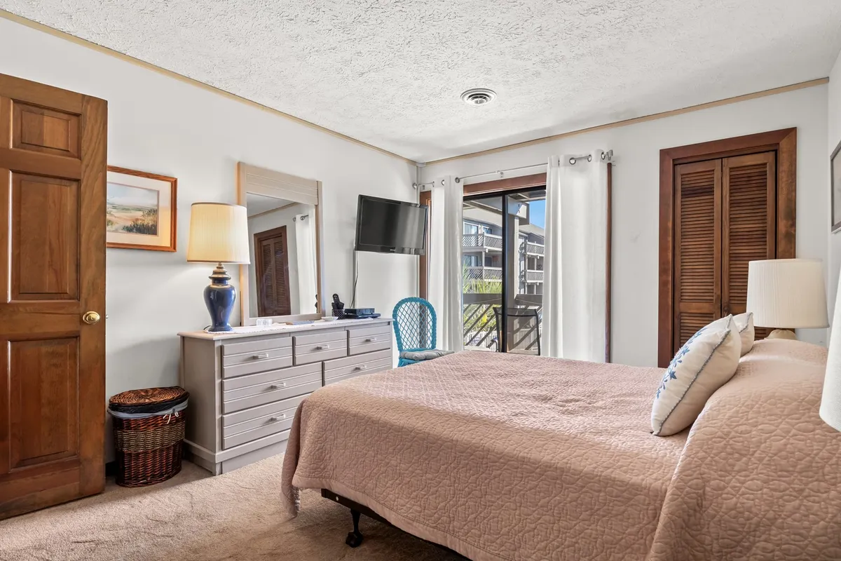 A cozy bedroom with a bed covered in a quilt, a dresser with a mirror, window with curtains, and a TV on a stand. The room feels welcoming and homelike, ideal for unwinding or enjoying private moments.