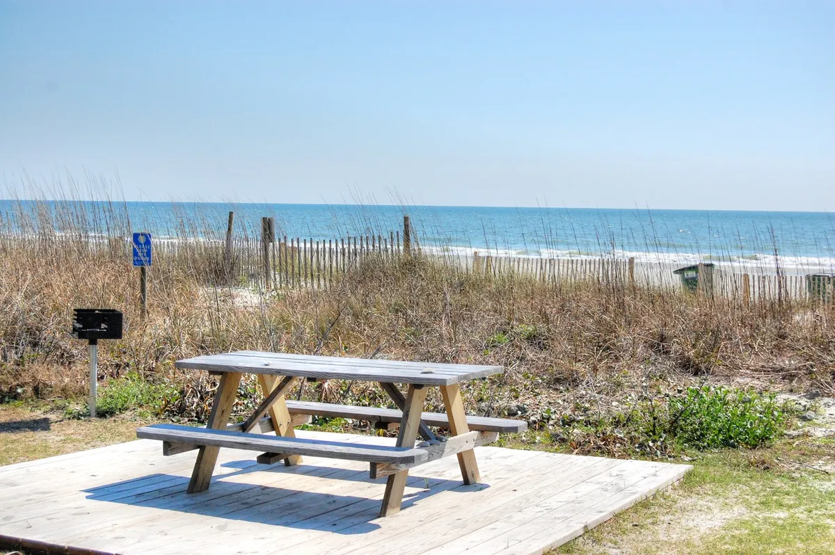A peaceful picnic table set atop dune grass, overlooking the ocean beyond a rustic fence. The spot offers a scenic place for meals, barbecues, or simply enjoying the sea breeze with loved ones.