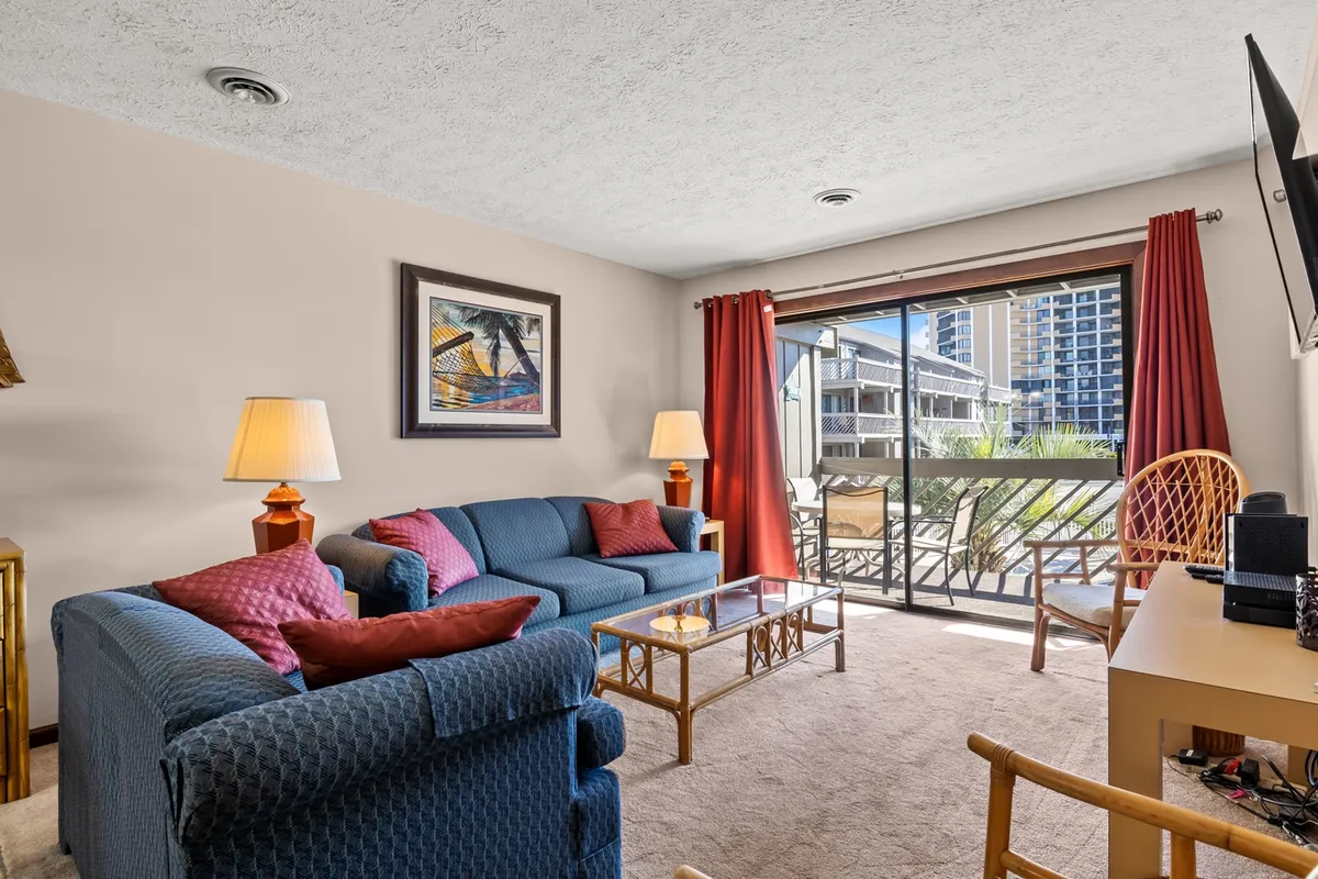 The image showcases a cozy condo living area featuring a comfortable sofa, modern coffee table, large sunlit window perfect for morning coffee, and cheerful artwork on the walls. The space feels bright, inviting, and ideal for relaxing or socializing with family and friends.