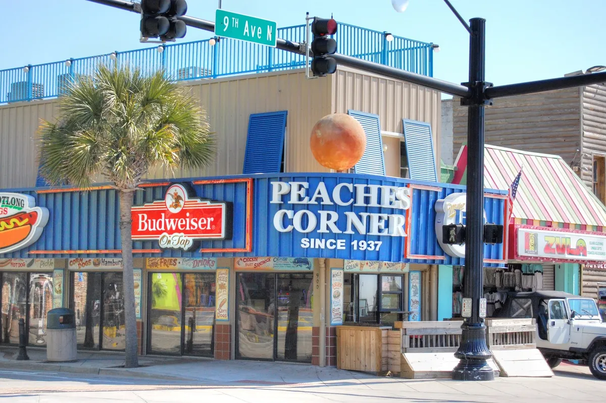 A festive exterior shot highlighting the iconic Peaches Corner and Budweiser signage, framed by palm trees and colorful storefront awnings. The scene buzzes with energy, representing the rich local culture and vibrant dining scene.