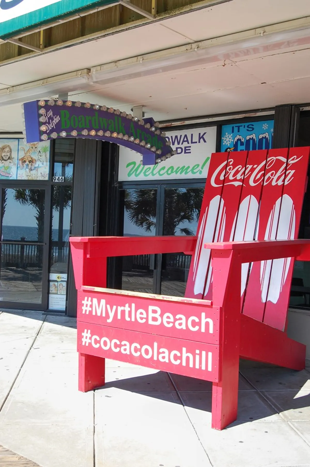 The playful scene features a large, colorful chair perfect for fun photo ops, with the Boardwalk Arcade sign and classic Coca-Cola branding in the background. The area feels lively and social, capturing the carefree mood of vacationers enjoying Myrtle Beach together.