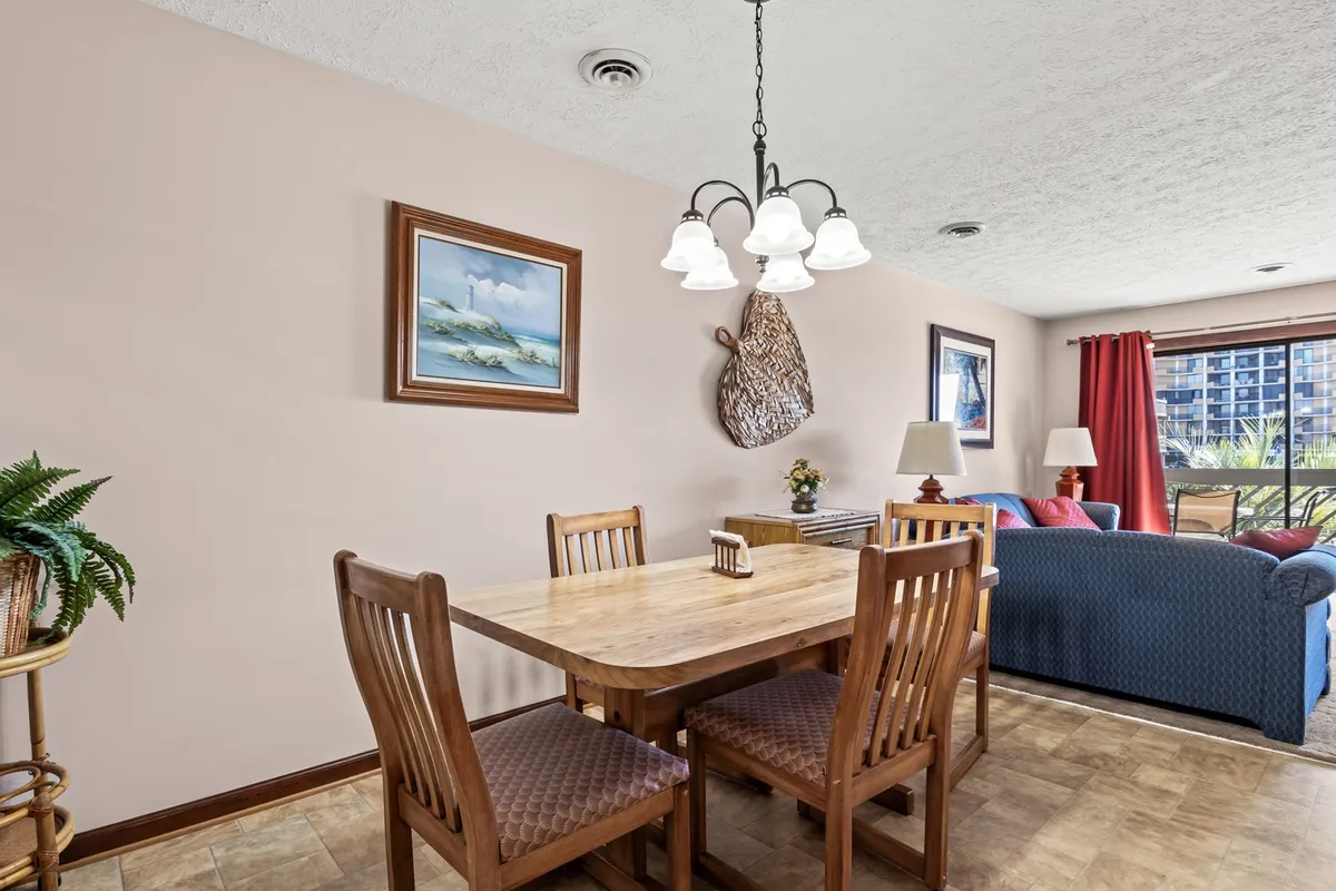 An open-plan dining area with a central dining table beneath a chandelier, artwork on the wall, and a view toward a comfortable living room seating area. The arrangement encourages shared meals and social interaction.