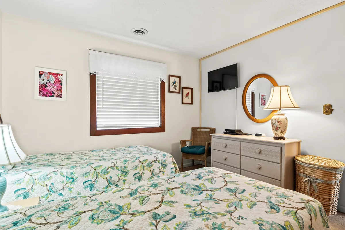 An inviting bedroom with two beds covered in floral quilts, a round mirror, a lamp, and framed artwork on the walls. The room exudes warmth, making it suitable for flexible sleeping arrangements for families or groups.
