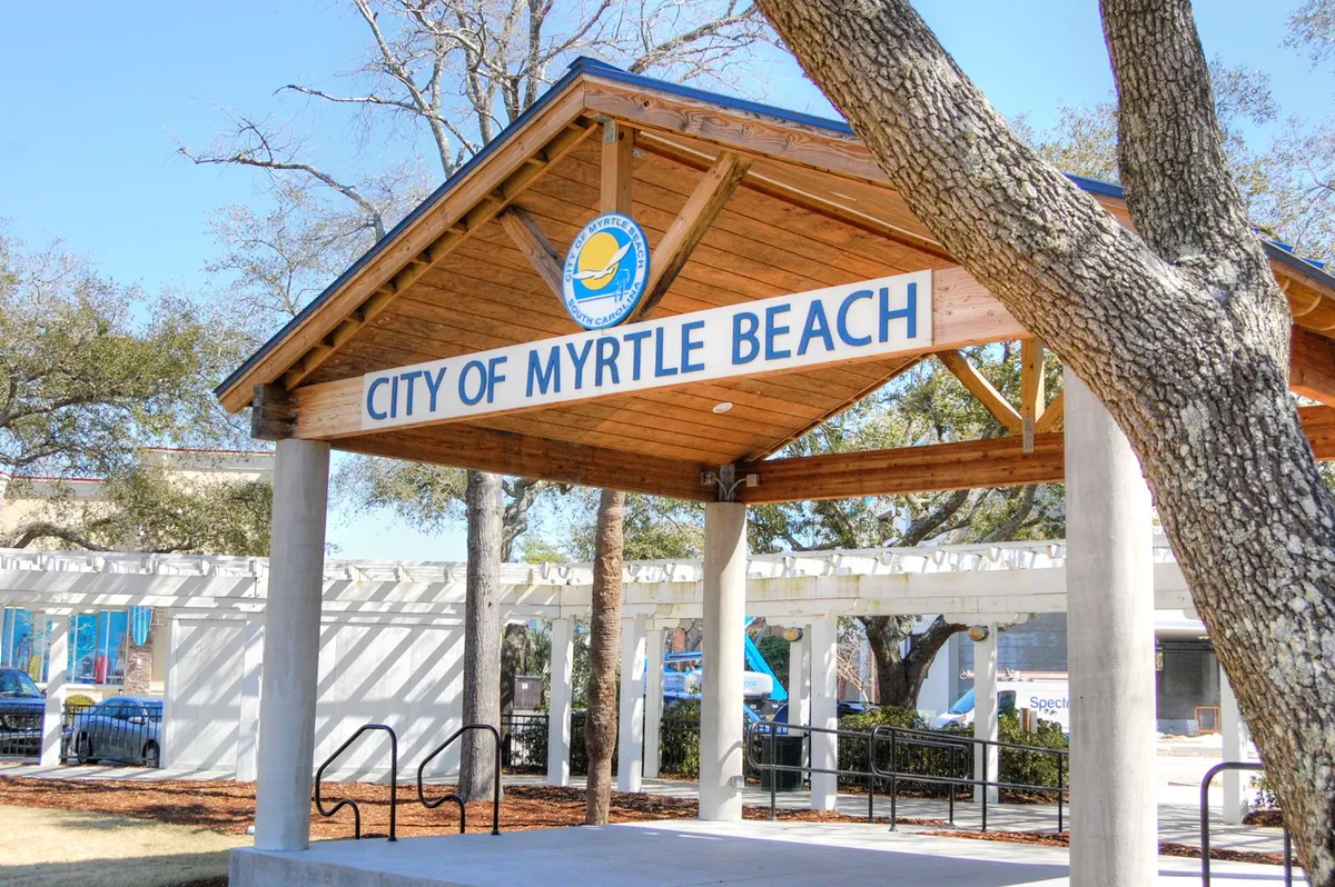 This image shows the welcoming signage for 'CITY OF MYRTLE BEACH' set against a backdrop of a wooden structure and leafy trees. The scene evokes a sense of arrival and highlights the destination's community atmosphere.