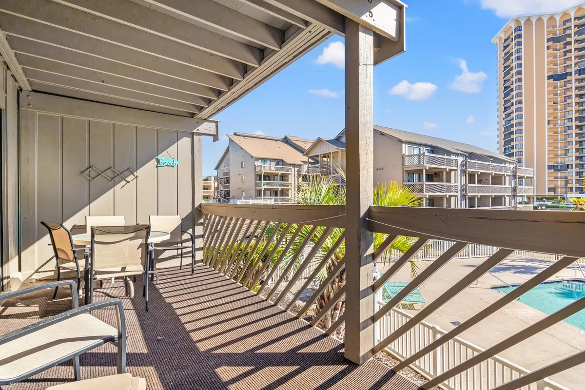 This image displays a balcony with a clear view of the pool area and surrounding resort buildings. The balcony railing and outdoor seating suggest an inviting spot to relax, enjoy a beverage, or chat with friends while overlooking the lively resort amenities below. The lighting hints at a time near sunset, adding warmth to the scene.