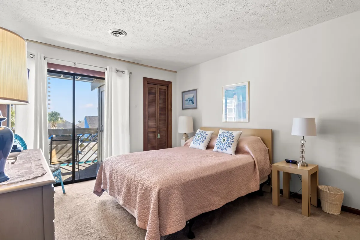 A bright and tranquil bedroom featuring a bed with a pink quilt, decorative pillows, and a lamp on the bedside table. Natural light streams in through a sliding glass door, giving the room a calm, homey feel.