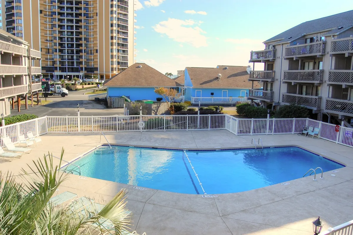 This image captures the swimming pool surrounded by the building and lush palm leaves, providing a clear view of the communal pool area where guests can swim, sunbathe, or gather for social activities. The relaxed, inviting mood is enhanced by the presence of poolside seating and the suggestion of group-friendly spaces.