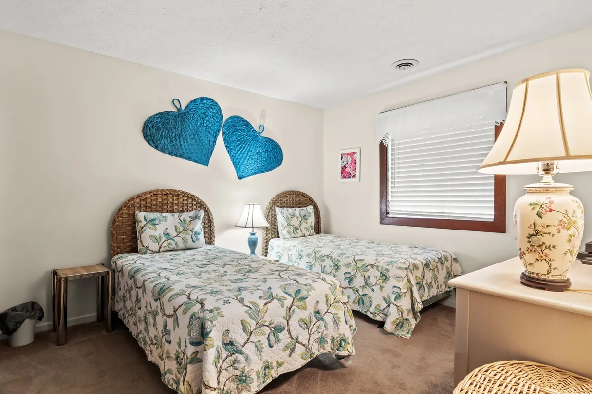 A charming twin bedroom with two twin beds adorned in floral bedding, blue heart-shaped wall decor above the beds, and matching lamps on each nightstand. The room feels bright and cheerful, perfect for kids or siblings to share.